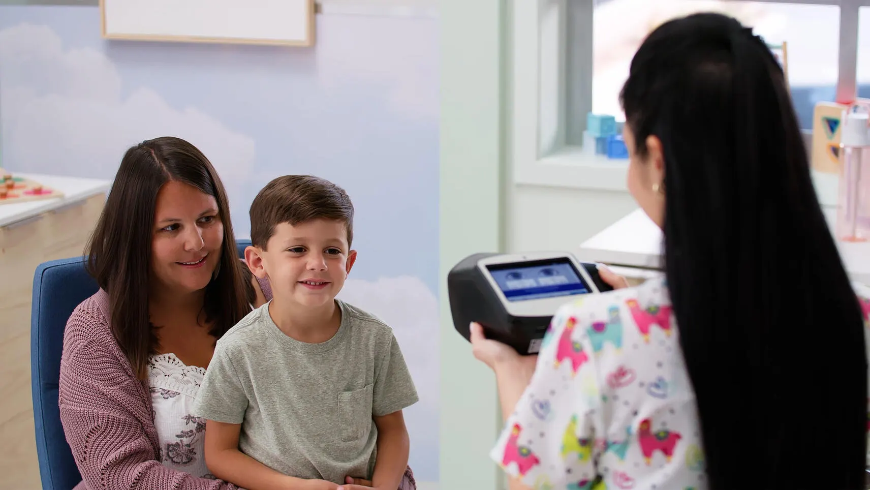 Healthcare worker in llama patterned scrubs points the Spot vision screener at a child in their mother's lap