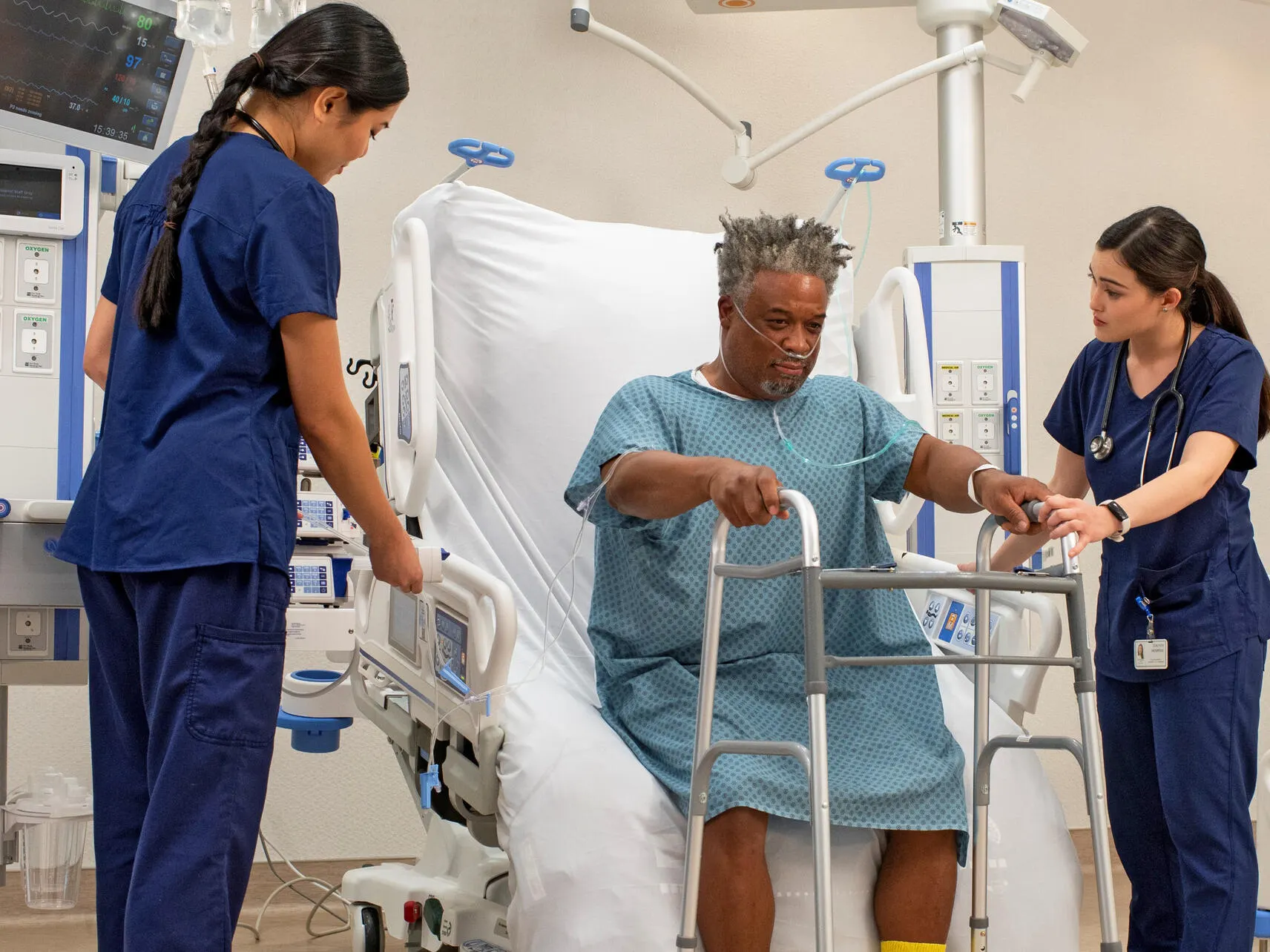 An elderly ICU patient being assisted to stand up by two healthcare professionals and a Progressa bed in a sitting position