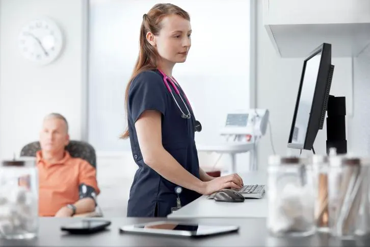 A healthcare worker enters information into a computer next to a sitting patient who is having their blood pressure checked