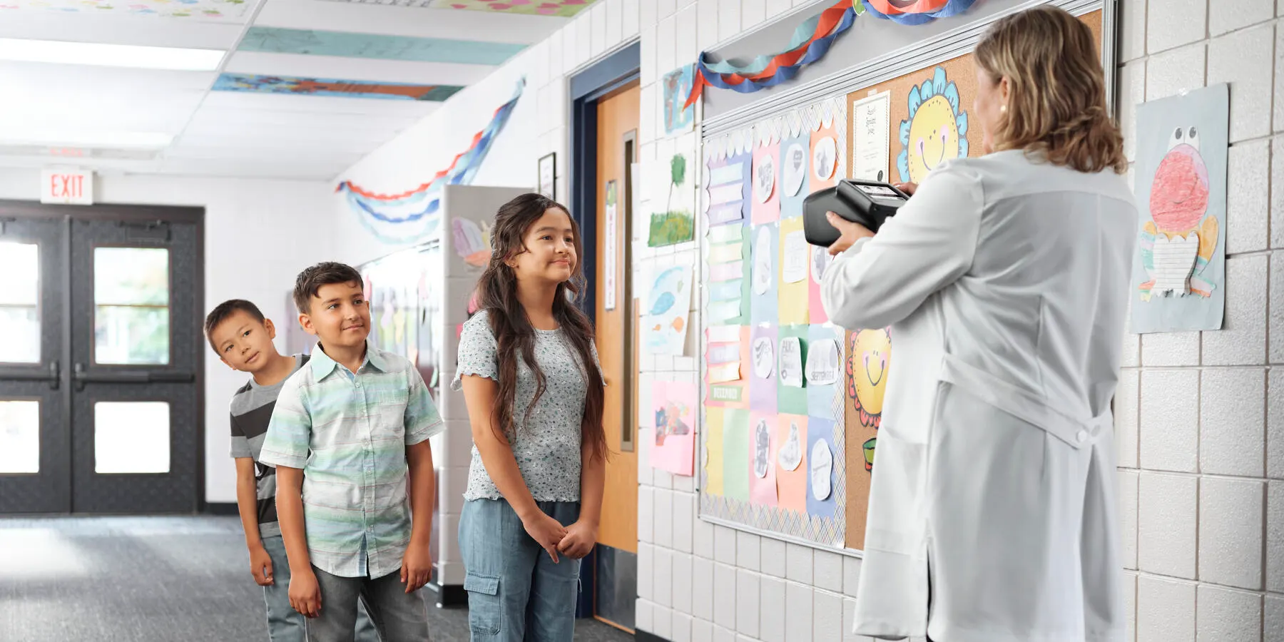 Healthcare worker in a lab coat points the Spot vision screener towards a line of children in a school corridor