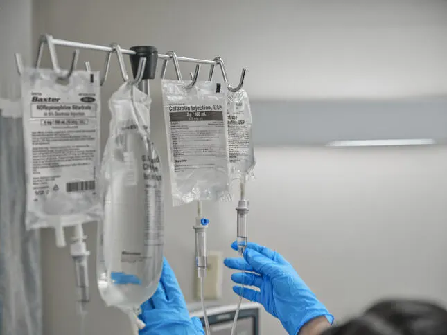 Gloved hands reaching for hanging IV bags of Cefazolin