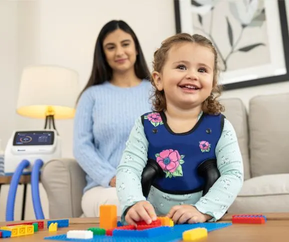 Child playing with legos while wearing the Vest APX, as her mom watches from behind.