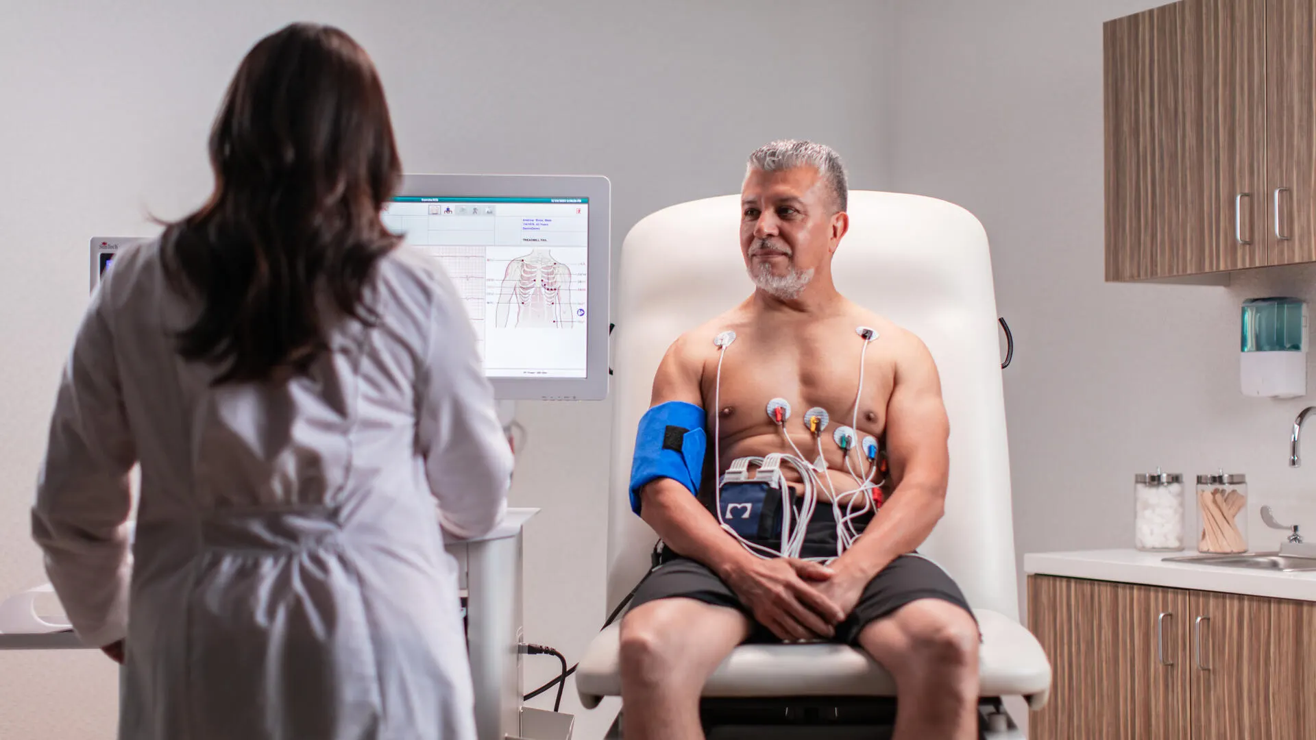 Cariology Technician examines an ECG next to a patient sitting up with electrodes on his chest