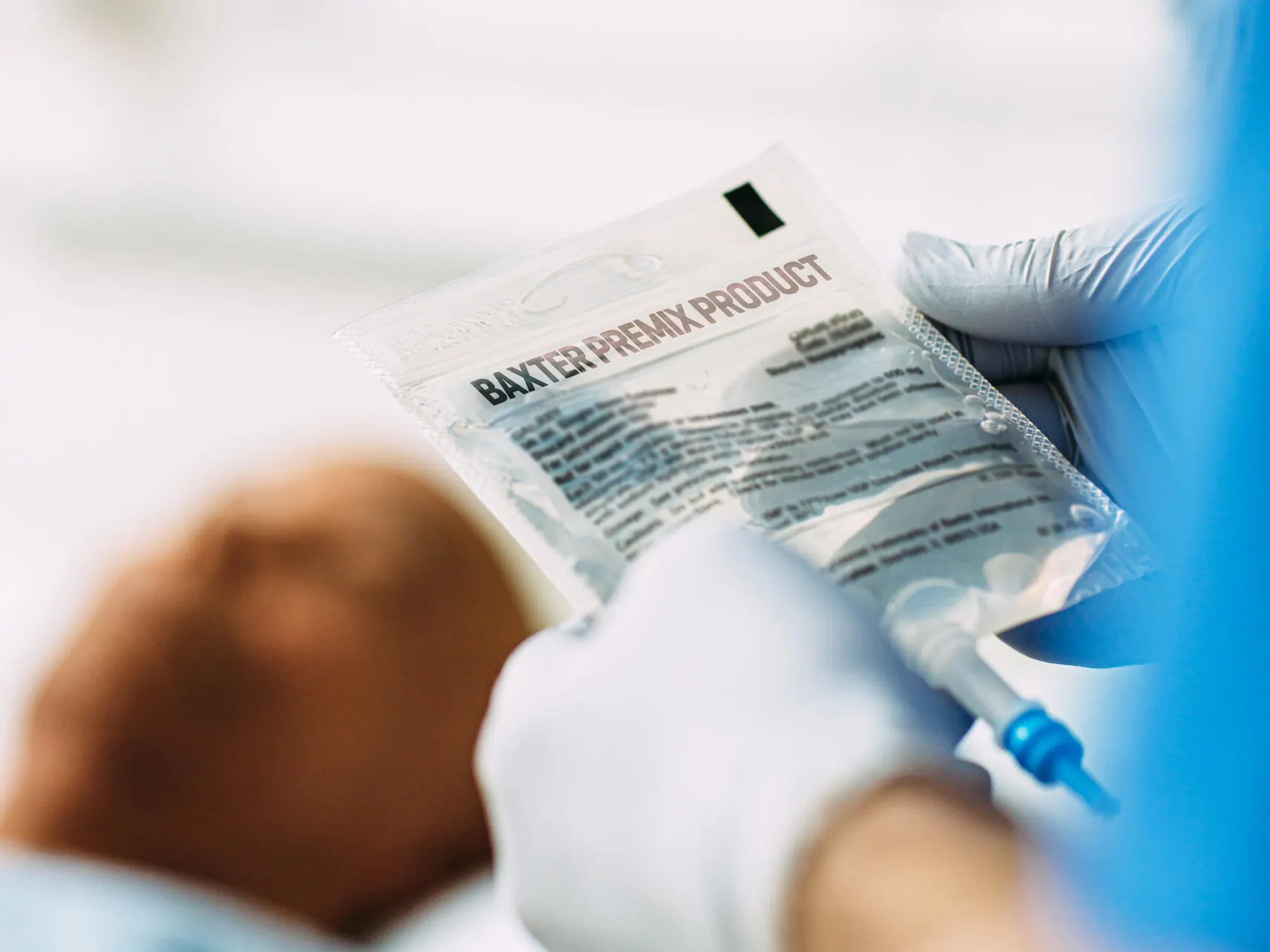 Gloved hands holding an IV bag of a Baxter premix product as a patient lies in a hospital bed in the background