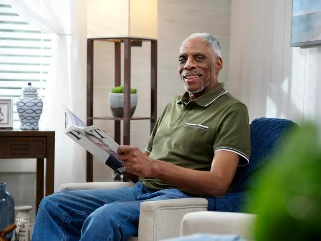 An elderly man sitting at home with a magazine, smiling at the camera.