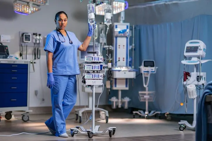 A healthcare worker poses in a hospital room next to infusion pumps with a bag attached