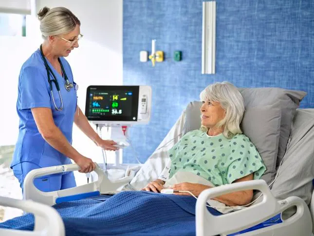 A healthcare worker talks to a patient attatched to a vital signs monitor in a hospital bed