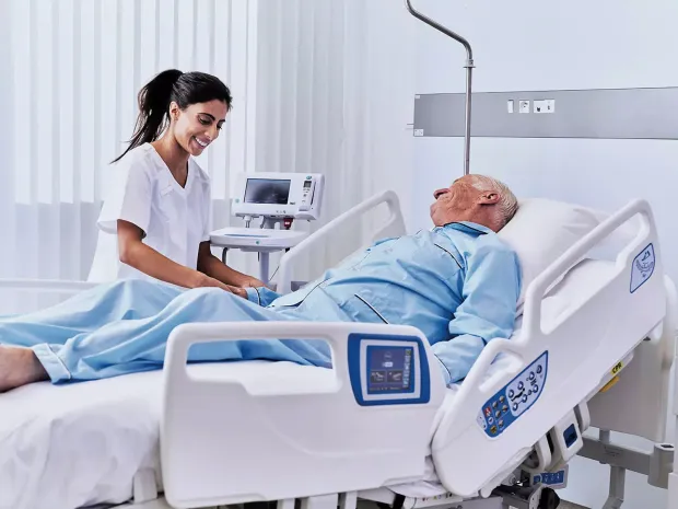 Caregiver assists a patient lying in pajamas on a hospital bed.