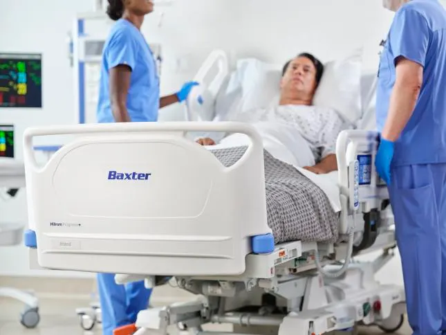 A patient in a Baxter hospital bed next to two healthcare workers in scrubs