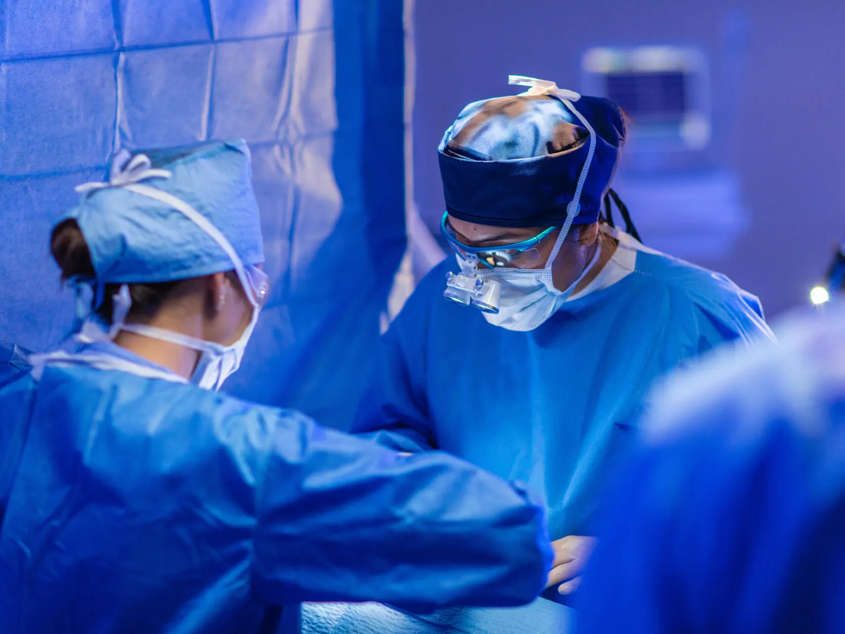 A surgeon wearing scrubs and loupes looks down at a patient