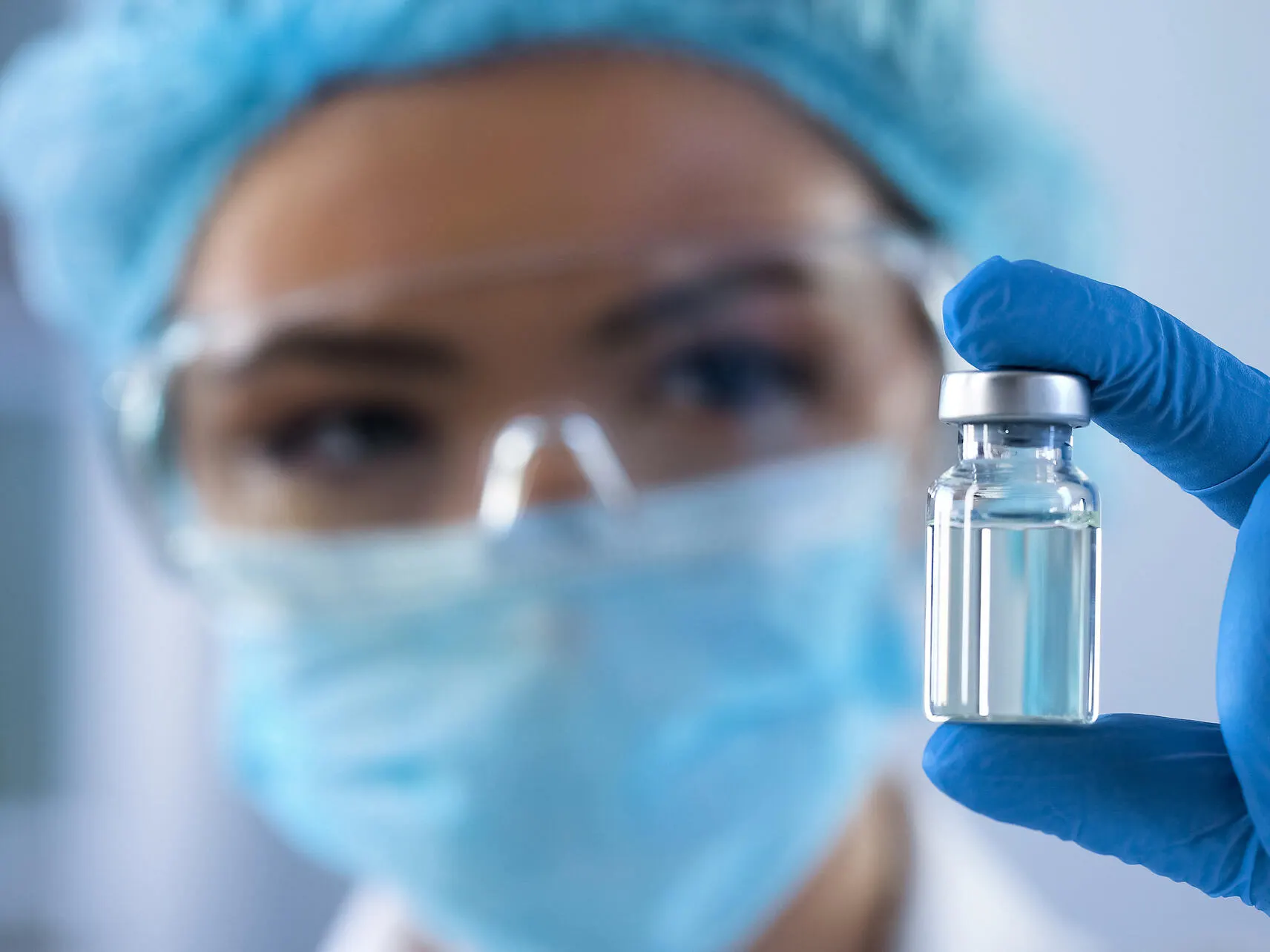 A clinician wearing a surgical mask, protective glasses, and blue gloves holds up a small glass vial containing a clear liquid