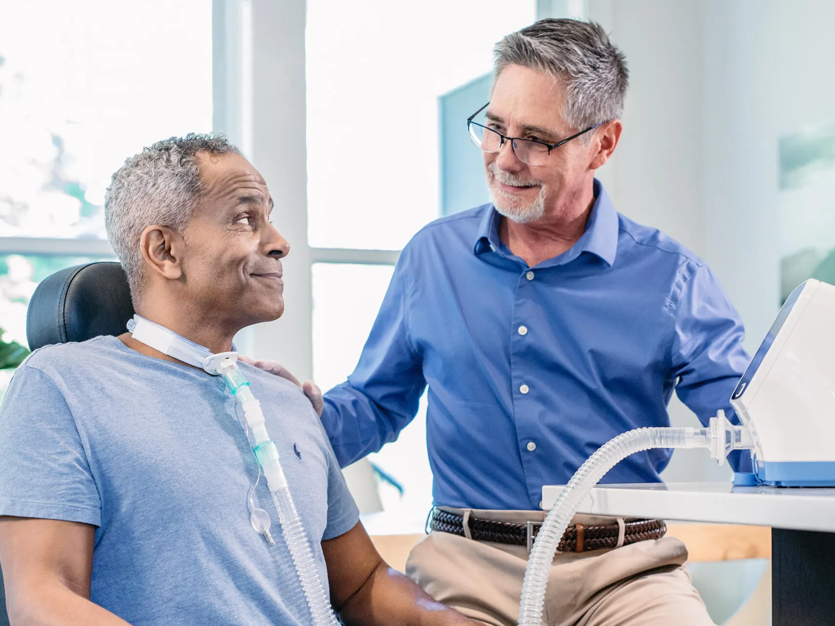 A patient trainer touches the shoulder of a patient receiving respiratory therapy via a tracheostomy port