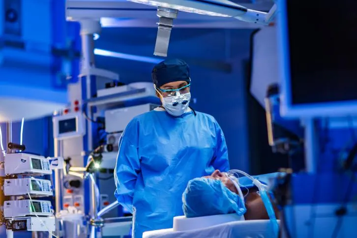 Surgeon stands over a patient on an operating table, surrounded by monitors and medical equipment.
