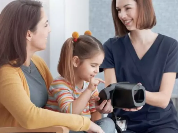 A smiling child sits with her mom while a nurse is showing her the Welch Allyn Spot Vision Screener device
