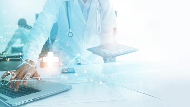 A doctor in a white coat types on a laptop while holding a tablet. Photo is overlaid with healthcare related graphics